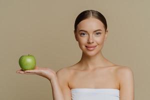 foto van gezonde, mooie vrouw met natuurlijke make-upstandaard gewikkeld in badhanddoek houdt groene appel aan beveelt product voor diëten aan geïsoleerd op bruine achtergrond maakt natuurlijke zelfgemaakte gezichtsmaskers