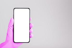 een hand- in een roze medisch handschoen houdt een zwart smartphone met een blanco wit scherm en geïsoleerd Aan een grijs achtergrond. foto