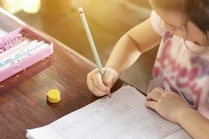 Aziatisch meisje zittend Aan een bureau Bij huis schrijven een boek. online onderwijs. nieuw normaal onderwijs. kinderen lezing en schrijven concepten. onderwijs van huis foto