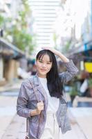 zelfverzekerd jong Aziatisch vrouw wie draagt een bruin gestreept blazer en schouder zak glimlacht terwijl ze zetten haar hand- Aan hoofd wandelen en glimlachen buitenshuis in de stad. foto