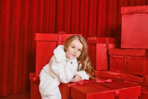 schattig weinig meisje in wit jas door de reusachtig rood Kerstmis presenteert dozen Aan rood achtergrond. vakantie, winkelen, plezier, mode foto