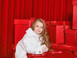 schattig weinig meisje in wit jas door de reusachtig rood Kerstmis presenteert dozen Aan rood achtergrond. vakantie, winkelen, plezier, mode foto
