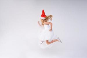 blij mooi meisje in rood de kerstman claus hoed en wit jurk lacht en springt foto