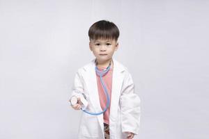 jong schattig Aziatisch jongen vervelend een medisch uniform Holding een stethoscoop spelen gelukkig dokter Aan wit achtergrond. peuter- kinderen doen alsof naar worden een kinderarts. foto