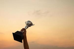 zege concept met een hand- Holding een trofee Aan de zonsondergang lucht achtergrond foto