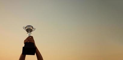 zege concept met een hand- Holding een trofee Aan de zonsondergang lucht achtergrond, banier ontwerp foto