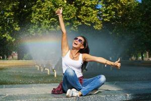 vrolijk jong vrouw in zonnebril zit tegen backdrop fontein met regenboog foto