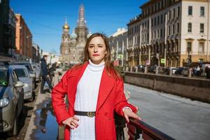 een jong vrouw in een rood jas wandelingen in de omgeving van de stad Aan een zonnig dag poseren voor de camera foto