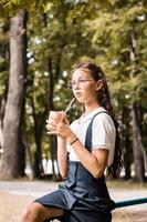 een schoolmeisje in bril drankjes een drank van een eco papier kop met een rietje in de park foto