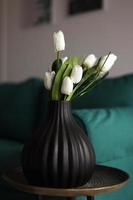 kunstmatig wit tulpen en groen bladeren in een zwart vaas Aan de tafel in huis interieur.
