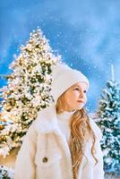 schattig Kaukasisch blond meisje in een witte ecobontjas, muts en handschoenen die in het winterkerstbos wandelen. nieuwjaar, sprookje, modeconcept foto