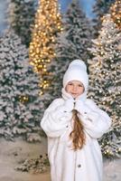 schattig Kaukasisch blond meisje in een witte ecobontjas, muts en handschoenen die in het winterkerstbos wandelen. nieuwjaar, sprookje, modeconcept foto