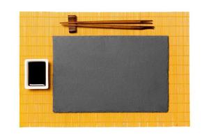 leeg rechthoekig zwart leisteen bord met eetstokjes voor sushi en soja saus Aan geel bamboe mat achtergrond. top visie met kopiëren ruimte voor u ontwerp foto