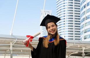glimlachen vrouw in diploma uitreiking japon Holding academisch mate en op zoek Bij camera met gebouw achtergrond foto