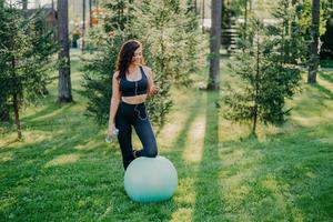 buitenopname van sportieve jonge vrouw houdt fles vers water vast, luistert naar muziek in oortelefoons, houdt moderne smartphone vast, leunt knie op fitnessbal, geniet van favoriete afspeellijst, poseert in groen bos foto