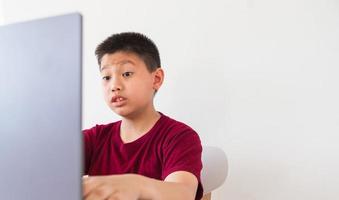 Aziatisch schattig weinig jongen is pratend online met zijn vrienden gelukkig kind maakt grappig gezichten geschokt en opgewonden Bij de laptop computer foto