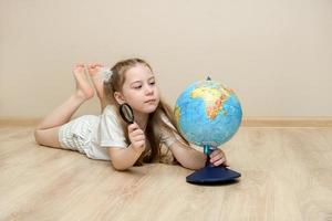 weinig schattig meisje leugens Aan de vloer, houdt een vergroten glas in een hand, en houdt de wereldbol in drie, voorzichtig aan het studeren het foto