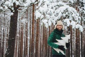 portret van knappe bebaarde man draagt warme bontmuts met oorkleppen en groene anorak, houdt kunstmatige dennenboom vast, poseert tegen hoge bomen bedekt met sneeuw, brengt vrije tijd door in winterbos foto