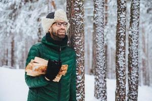 aangenaam uitziende gelukkige man houdt brandhout vast, kijkt peinzend opzij, staat in de buurt van winterbomen, droomt over iets aangenaams, geniet van prachtige winterlandschappen. natuur concept. foto