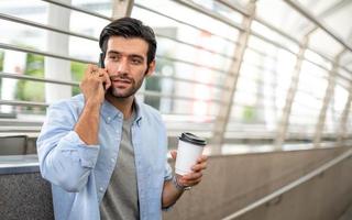 de Mens Holding een kop van koffie en gebruik makend van een smartphone en pratend met zijn vriend terwijl hij aan het wachten zijn vriend Bij de luchthaven. foto