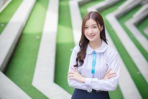 mooi hoog school- Aziatisch leerling meisje in de school- uniform staat en glimlacht gelukkig met een beugel Aan haar tanden terwijl ze arm kruispunt vol vertrouwen terwijl ze looks Bij de camera. foto