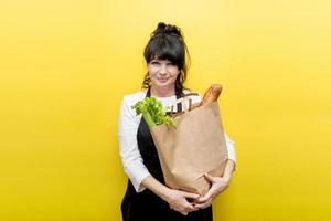 mooi ouderen dame koken in een zwart schort met een papier boodschappen doen zak met een baguette en selderij, de concept van werkzaamheid in oud leeftijd, Gezondheid, professionaliteit foto