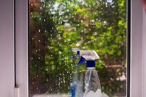 vuil venster nat gemaakt met schuim met een fles van wasmiddel. achter glas een boom met groen bladeren foto