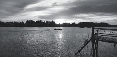 silhouet van houten waterkant, waterkant of brug met lange staart boot Aan rive en boom achtergrond Bij zonsondergang of zonneschijn tijd. mooi natuur en vervoer in zwart en wit toon of monochroom. foto