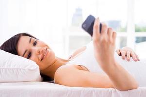 controle berichten van hem. mooi jong vrouw op zoek Bij haar mobiel telefoon en glimlachen terwijl aan het liegen in bed foto