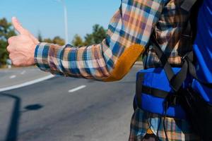 nu is tijd naar nemen een rijden. detailopname van jong Mens draag- rugzak uitrekken uit hand- met duim omhoog terwijl staand Aan de weg foto