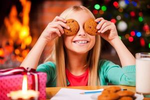 Kerstmis plezier. schattig weinig meisje aan het bedekken haar ogen met koekjes en glimlachen terwijl zittend Bij de tafel met Kerstmis boom en haard in de achtergrond foto