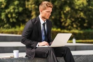 werken buitenshuis. zelfverzekerd jong Mens in overhemd en stropdas werken Aan laptop terwijl zittend buitenshuis foto