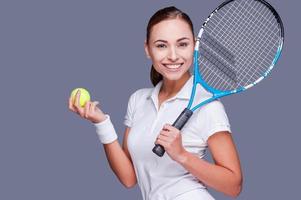 toetreden me voor tennis mooi jong Dames in sport- kleren Holding tennis racket Aan haar schouder en glimlachen terwijl staand tegen grijs achtergrond foto