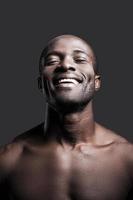 openhartig geluk. portret van jong zonder shirt Afrikaanse Mens houden ogen Gesloten en glimlachen terwijl staand tegen grijs achtergrond foto