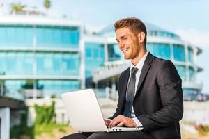 werken met genoegen. knap jong Mens in formele kleding werken Aan laptop en glimlachen terwijl zittend buitenshuis en tegen gebouw structuur foto