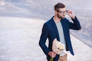 is dat haar knap jong Mens in slim gewoontjes slijtage Holding boeket van bloemen en aanpassen zijn bril terwijl wandelen door de straat foto