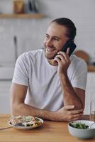 knap jong Mens pratend Aan mobiel telefoon en glimlachen terwijl hebben lunch Bij de keuken foto