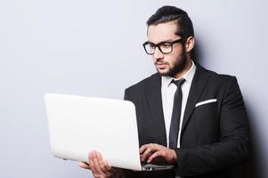 zakenman met laptop. zelfverzekerd jong Mens in formele kleding werken Aan laptop terwijl staand tegen grijs achtergrond foto