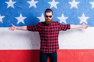 compleet vertrouwen. knap jong gebaard Mens in zonnebril houden armen opening en op zoek Bij camera terwijl staand tegen Amerikaans vlag foto