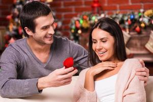 zullen u trouwen me knap jong Mens maken een voorstel terwijl geven een verloving ring naar zijn vriendin met Kerstmis decoratie in de achtergrond foto