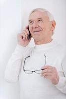 gebruik makend van modern gadgets. vrolijk senior Mens pratend Aan de mobiel telefoon en Holding bril terwijl leunend Bij de muur foto