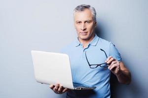 altijd klaar naar helpen jij. vrolijk senior Mens in gewoontjes Holding zijn bril en laptop terwijl staand tegen grijs achtergrond foto