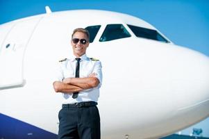 lucht is mijn passie. zelfverzekerd mannetje piloot in uniform houden armen gekruiste en glimlachen met vliegtuig in de achtergrond foto