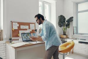 zelfverzekerd jong Mens op zoek Bij laptop terwijl staand in de buurt zijn werken plaats foto