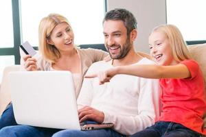 familie boodschappen doen online. gelukkig familie van drie bonding naar elk andere en glimlachen terwijl zittend Aan de bankstel en boodschappen doen online samen foto