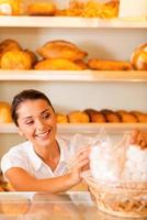 gelukkig bakker Bij werk. aantrekkelijk jong vrouw in schort inpakken koekjes en glimlachen terwijl staand in bakkerij winkel foto