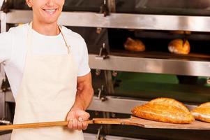 vers gebakken brood voor jij. bijgesneden beeld van zelfverzekerd jong Mens in schort nemen vers gebakken brood van oven en glimlachen foto