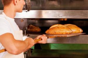 alleen maar gebakken. zelfverzekerd jong Mens in schort nemen de vers gebakken brood van oven foto