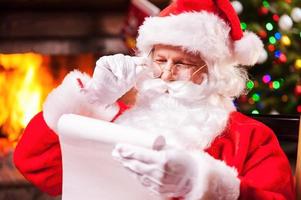 controle zijn speciaal lijst. traditioneel de kerstman claus zittend Bij zijn stoel en lezing een brief met haard en Kerstmis boom in de achtergrond foto