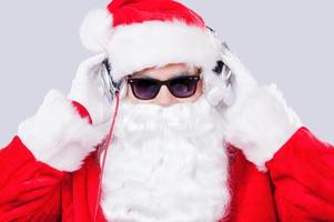 koel de kerstman. de kerstman claus in zonnebril aanpassen zijn hoofdtelefoons terwijl staand tegen grijs achtergrond foto